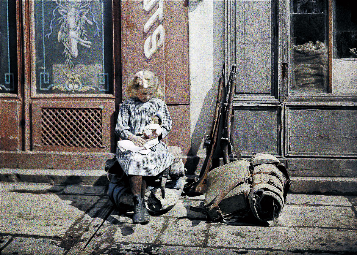 ایک چھوٹی بچی اپنی گڑیا کے ساتھ کھیل رہی ہے۔ اس کے پہلو میں دو بندوقیں اور ایک فوجی تھیلا زمین پر رکھا ہے۔ پہلی جنگ عظیم، مغربی محاذ، فرانس۔ 1917ء 